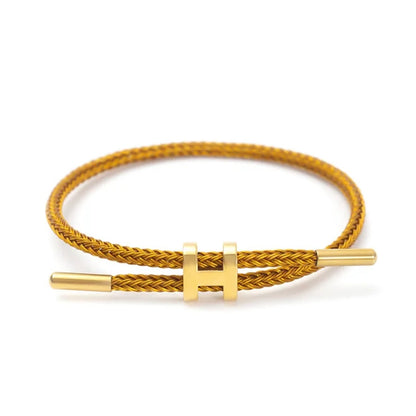 A product photo of a minimalist gold bracelet. The bracelet features a braided gold-colored cord and a sleek, adjustable gold metal clasp in the shape of a capital "H." The background is a clean white.