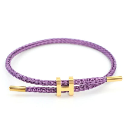 A studio photo of a minimalist purple bracelet. The bracelet is made of a braided purple rope and features a sleek gold-toned metal clasp in the shape of a capital 'H' that can be adjusted for size. The bracelet is displayed on a clean white background.