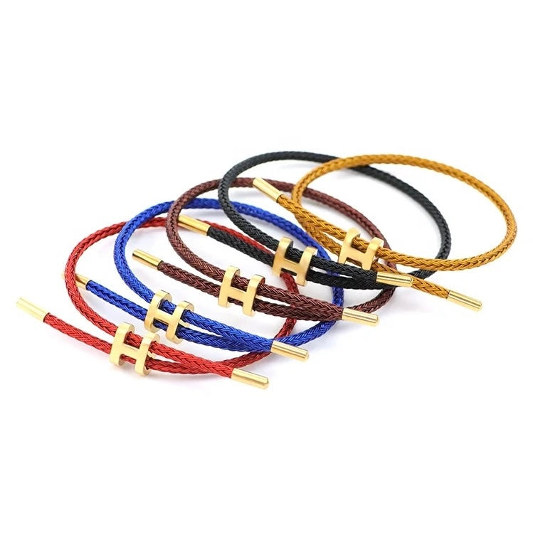 A product photo of five adjustable bracelets stacked on a white background. The bracelets have a minimalist design, with braided rope in various colors: red, blue, dark brown, black, and golden-yellow. Each bracelet features an adjustable gold-toned metal clasp in the shape of a capital 'H'.