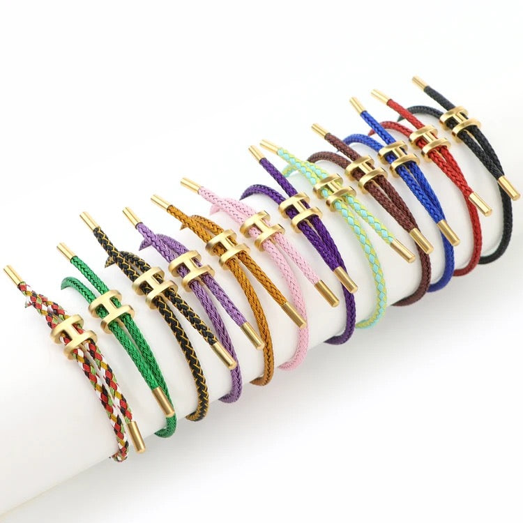 A wide-angle product photo showing a collection of twelve adjustable bracelets arranged side-by-side on a white surface. Each bracelet features a braided cord in a different color, including solid and multi-colored options. They all have a matching gold-toned metal clasp in the shape of a capital 'H'. The bracelets are displayed in a gentle curve on the surface.