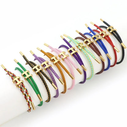 A wide-angle product photo showing a collection of twelve adjustable bracelets arranged side-by-side on a white surface. Each bracelet features a braided cord in a different color, including solid and multi-colored options. They all have a matching gold-toned metal clasp in the shape of a capital 'H'. The bracelets are displayed in a gentle curve on the surface.