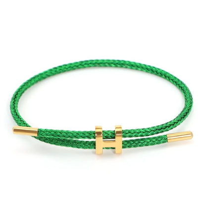 A high-resolution product shot of a minimalist bracelet. The bracelet is made from a vibrant green braided rope and features a shiny gold-colored metal clasp in the shape of a capital 'H'. The clasp is designed to slide, allowing the bracelet to be adjusted. The background is a clean, solid white.