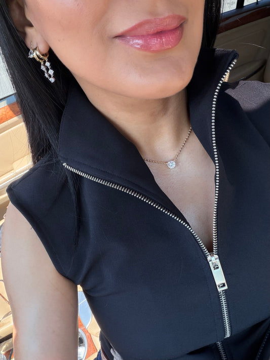 Woman in a dark zip top wearing a delicate necklace with a small clear stone at the collarbone