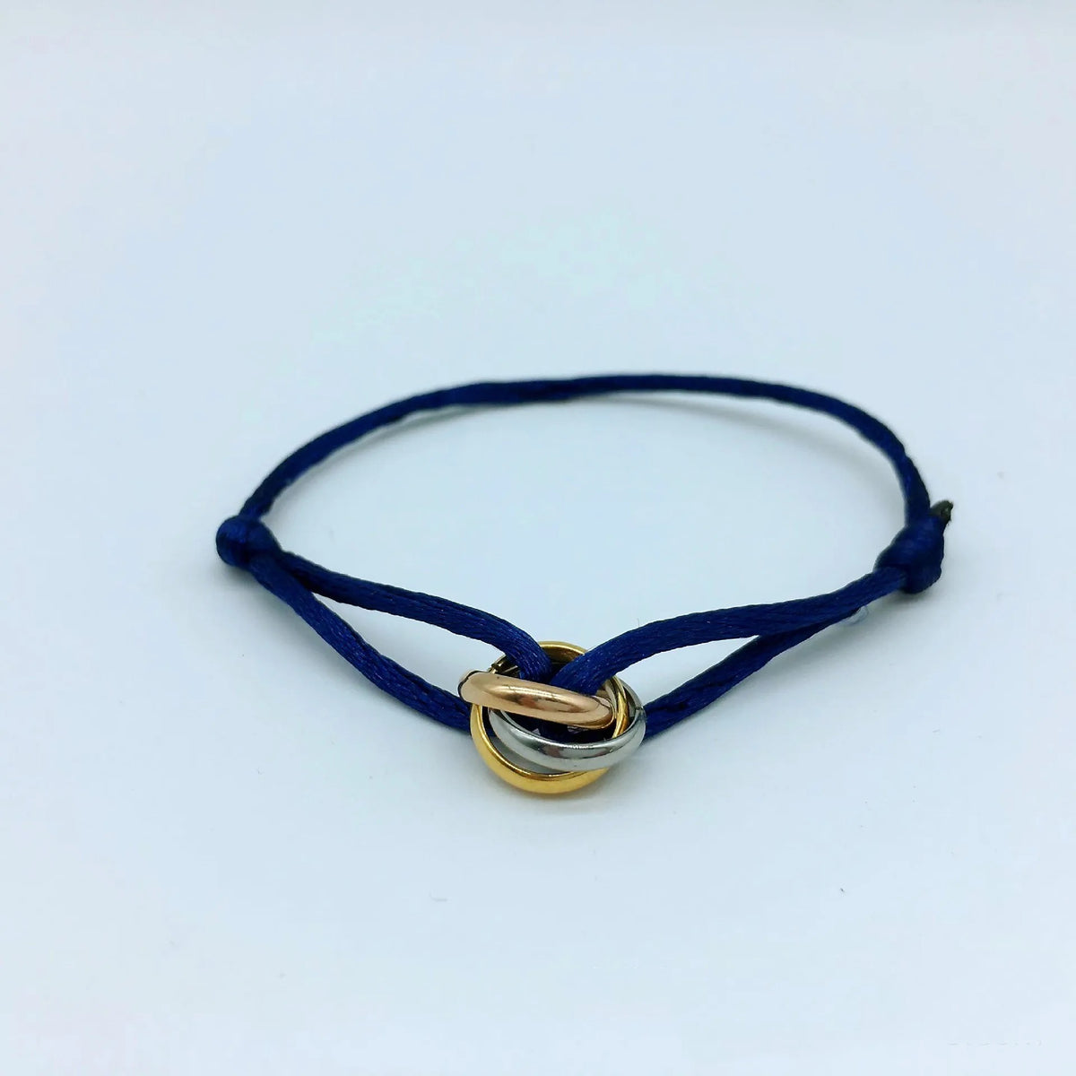 Pulsera de cordón azul marino con tres pequeños anillos entrelazados, sobre un fondo azul claro