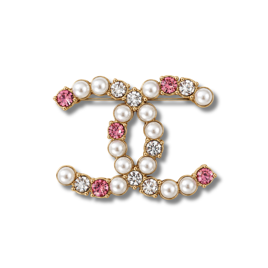 Broche tono dorado de doble curva entrelazada con perlas color crema y piedras transparentes y rosas, sobre fondo blanco