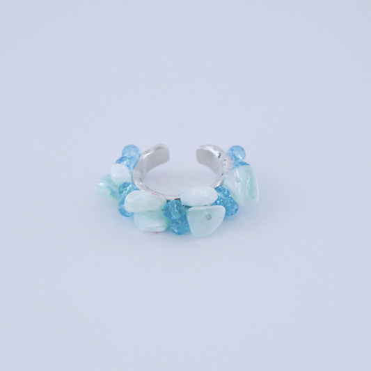 Anillo abierto con fragmentos de piedra azul claro y aguamarina sobre una banda en tono plateado, sobre una superficie azul pálida