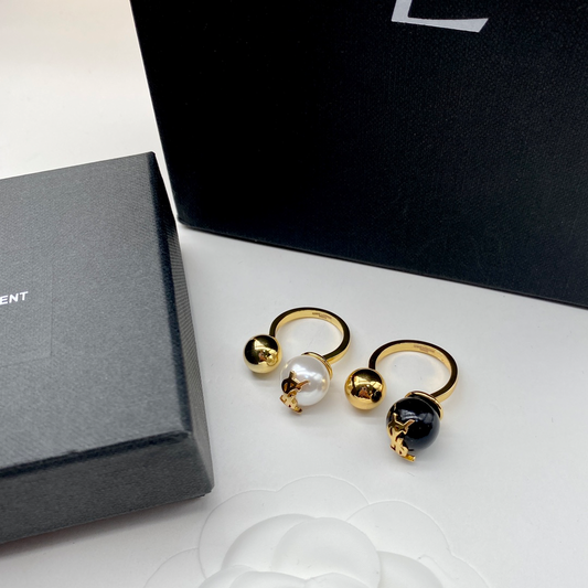 Dos anillos abiertos con una bola de color dorado y una cuenta redonda, uno blanco y uno negro, frente a una caja oscura