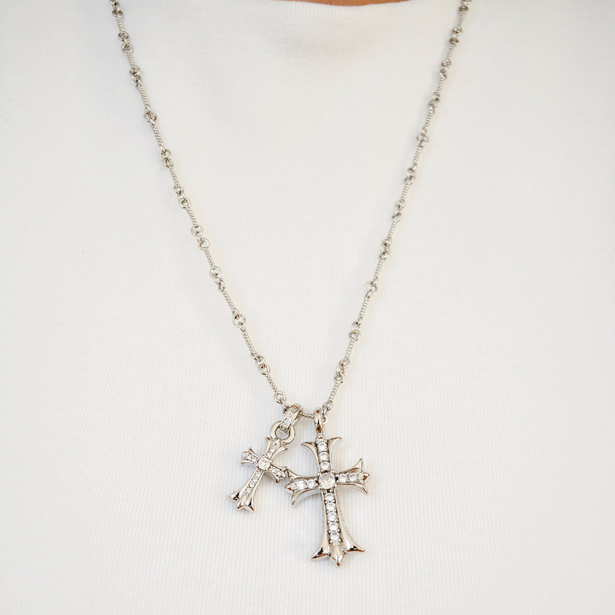 Collar de cadena en tono plateado con dos colgantes de cruz con piedras transparentes, colgando sobre una superficie clara