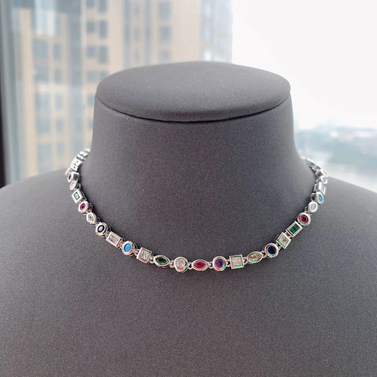 Collar en tono plateado con piedras de colores de formas variadas sobre un busto de exhibición gris