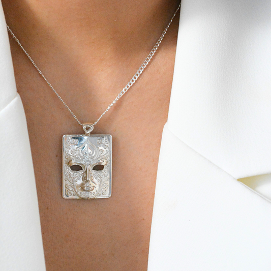Silver-tone rectangular plaque pendant with a masked face in relief, worn at the neck over a white blazer